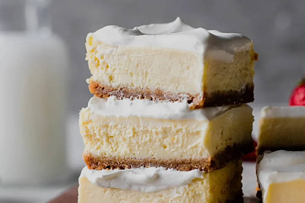 Three layers of creamy cheesecake bars with a graham cracker crust and topped with whipped cream, stacked on a wooden surface, with a glass of milk in the background.