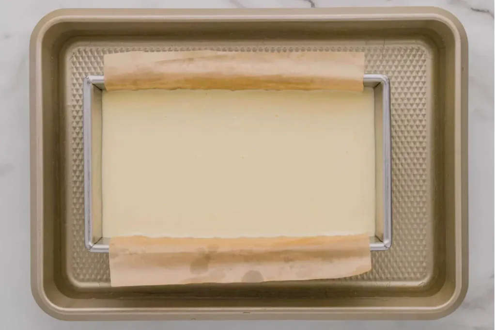 Overhead view of a baking tray with a wire rack and parchment strips framing a smooth, pale rectangular layer; a neat, quiet kitchen scene.