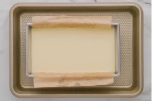 Overhead view of a baking tray with a wire rack and parchment strips framing a smooth, pale rectangular layer; a neat, quiet kitchen scene.