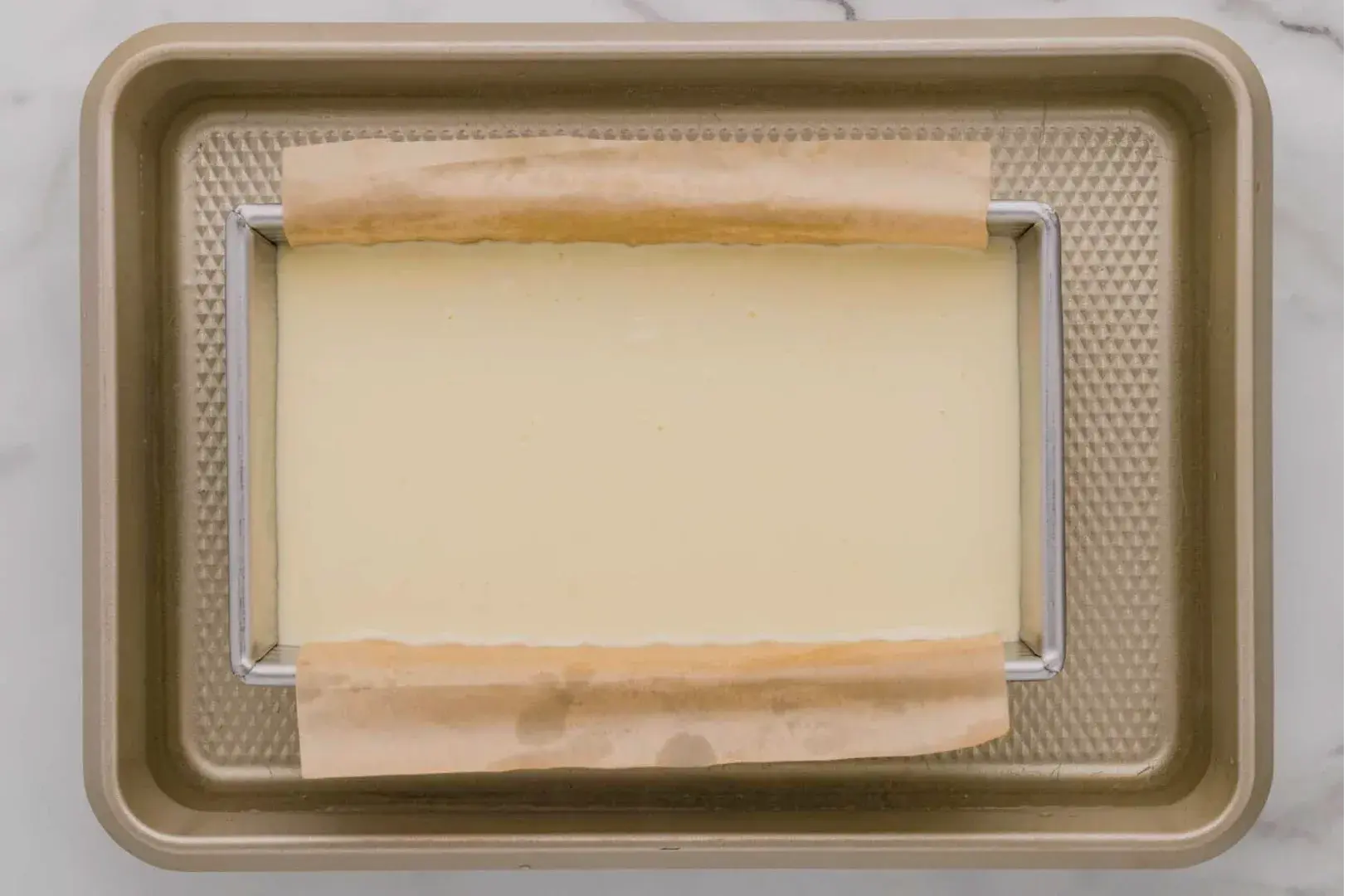 Overhead view of a baking tray with a wire rack and parchment strips framing a smooth, pale rectangular layer; a neat, quiet kitchen scene.