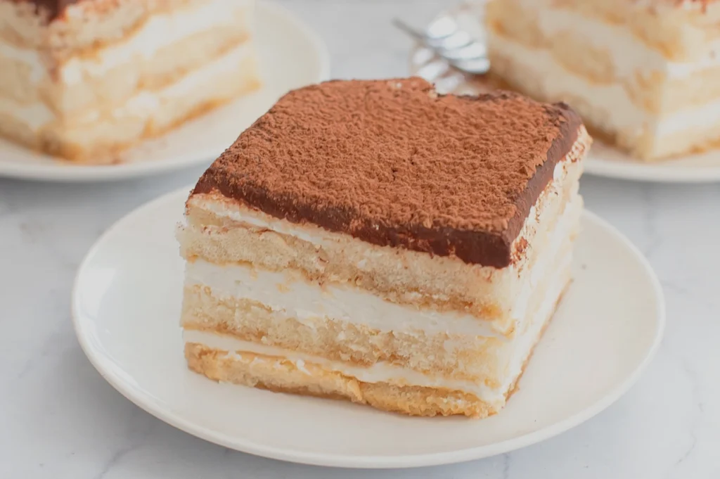 Slice of classic tiramisu layered with mascarpone cream and coffee-soaked sponge, dusted with cocoa on a white plate; cozy dessert setting.