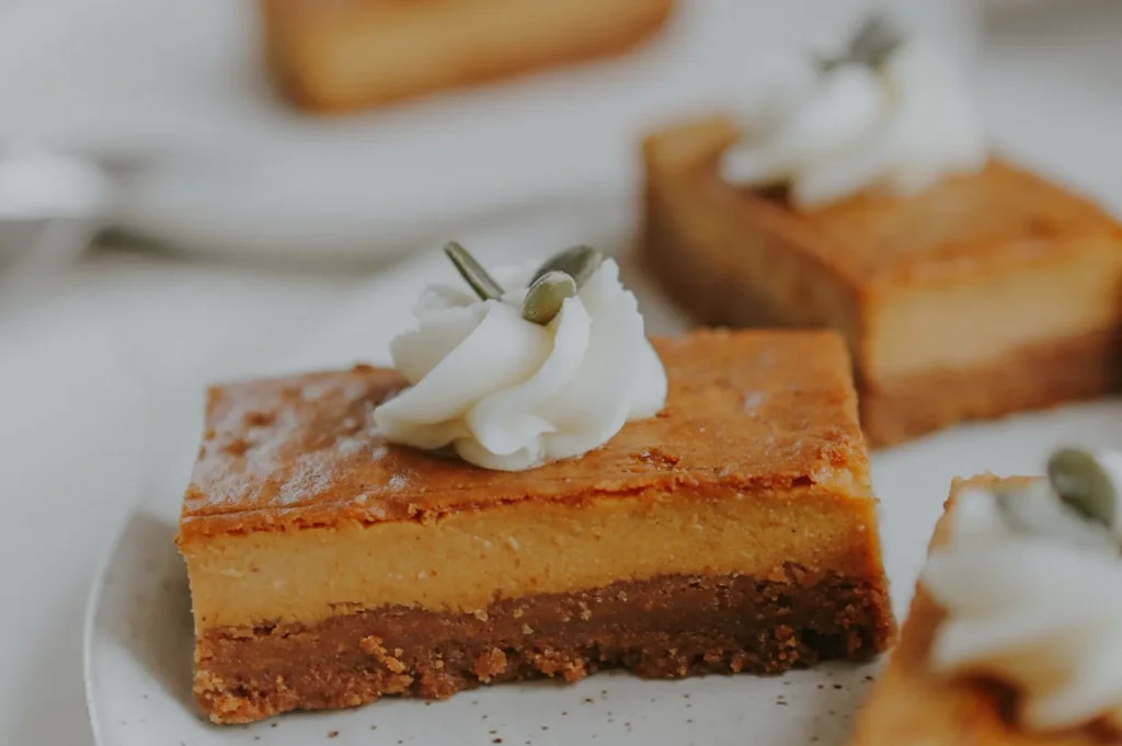 A close-up of dessert squares on a white plate, featuring a layer of creamy filling topped with a dollop of whipped cream and a pumpkin seed.