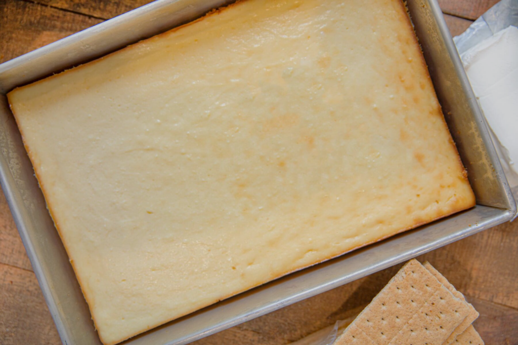 Golden-brown cheesecake in a rectangular pan on a wooden table, with graham crackers lying nearby, evoking a warm, homemade feel.