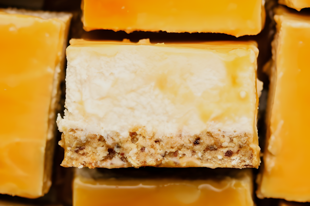 Close-up of a caramel cheesecake bar with a smooth caramel glaze, creamy cheese layer, and a nutty crust. The bars are arranged closely, creating a rich, indulgent feel.