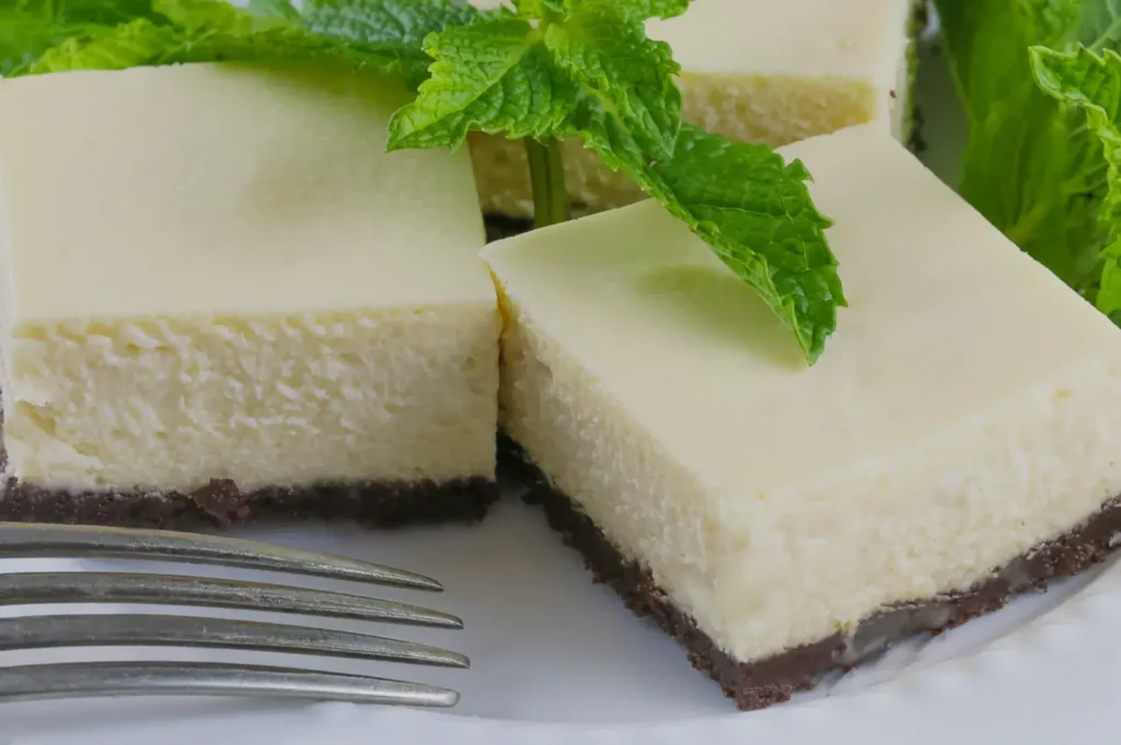 A close-up of creamy cheesecake squares with a chocolate crust, garnished with fresh mint leaves, served on a white plate with a fork.