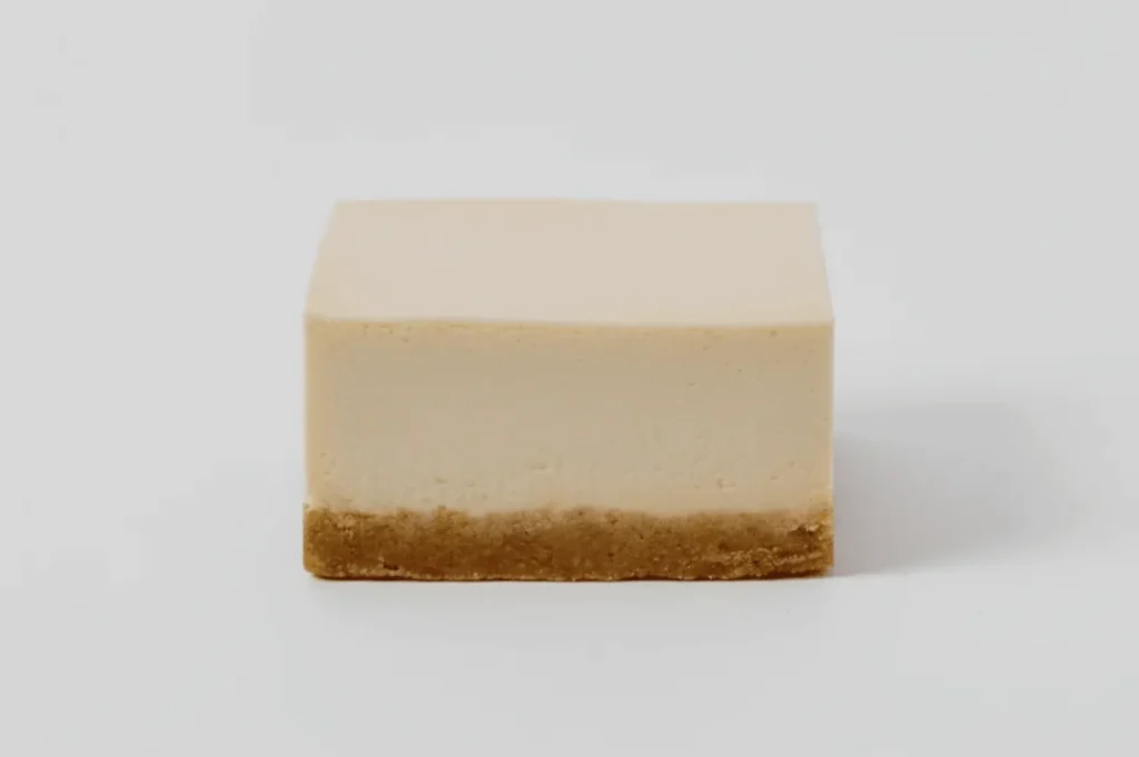 A neatly sliced rectangular cheesecake with a creamy beige layer and crumbly golden-brown crust, isolated on a white background, evoking simplicity.