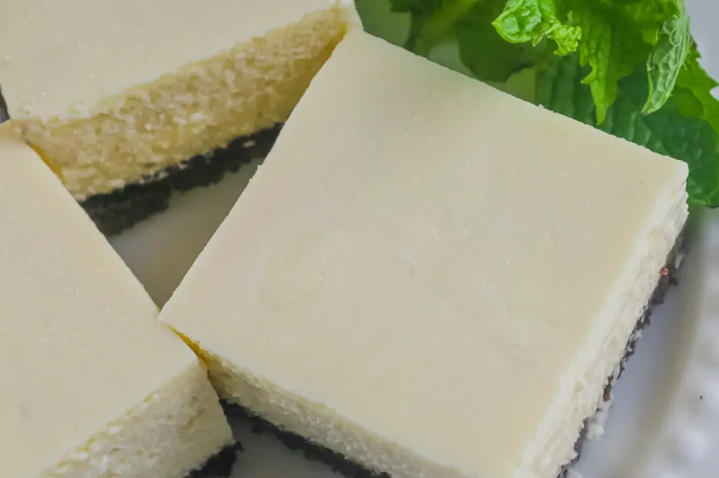 A square slice of creamy white cheesecake with a dark crumb crust sits on a plate beside two slices, garnished with fresh mint leaves.