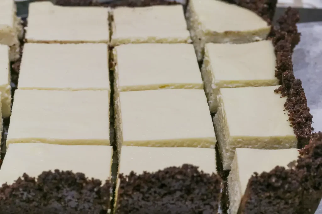 Neatly cut squares of creamy cheesecake with dark chocolate crumb crust are arranged in rows on a tray, creating an inviting dessert display.