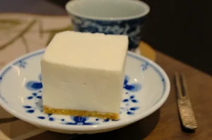 A square piece of dessert on a decorative blue and white plate, with a small bowl in the background. A silver fork is beside the plate.
