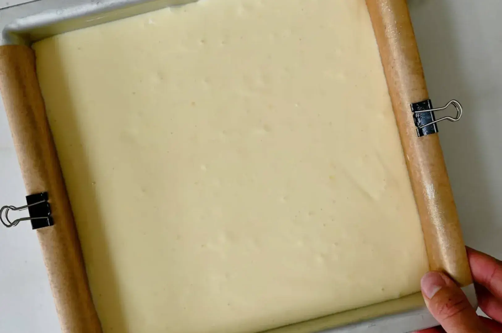 A baking pan with a smooth, creamy batter spread evenly inside, and two wooden dowels attached to the sides with clips.