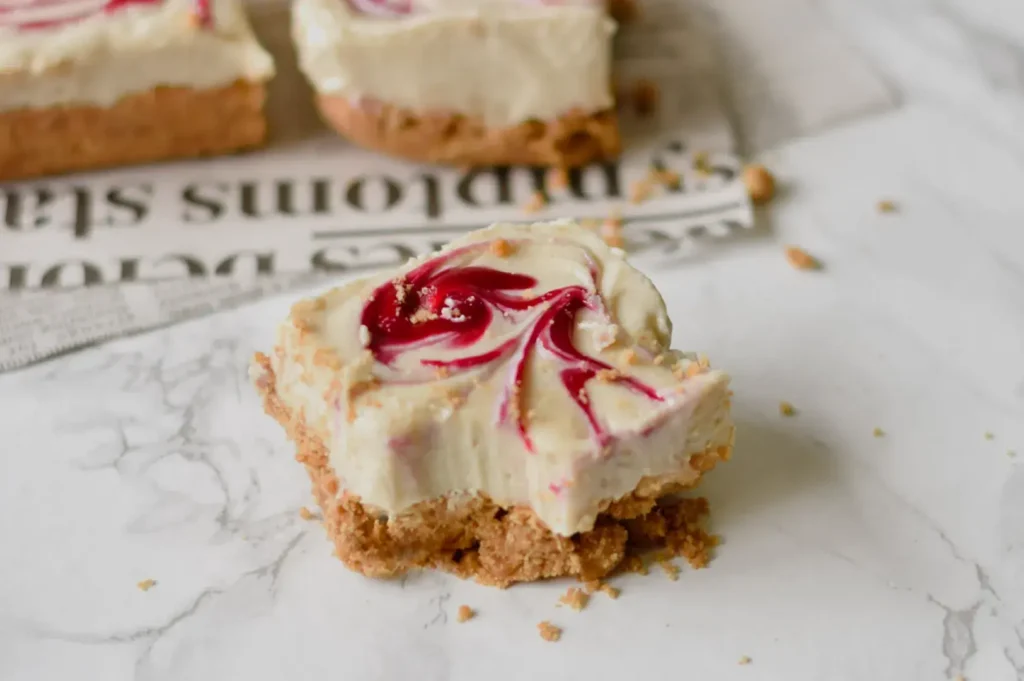 A slice of cheesecake with a raspberry swirl on a crumbly crust, placed on a marble surface.
