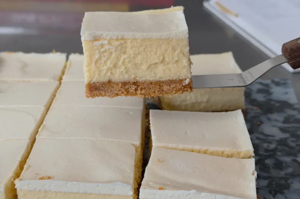 A slice of cheesecake being lifted from a tray, with the rest of the cheesecake cut into squares in the background.