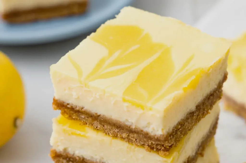 A stack of lemon cheesecake bars with a graham cracker crust, topped with a yellow lemon swirl, with a lemon in the background.