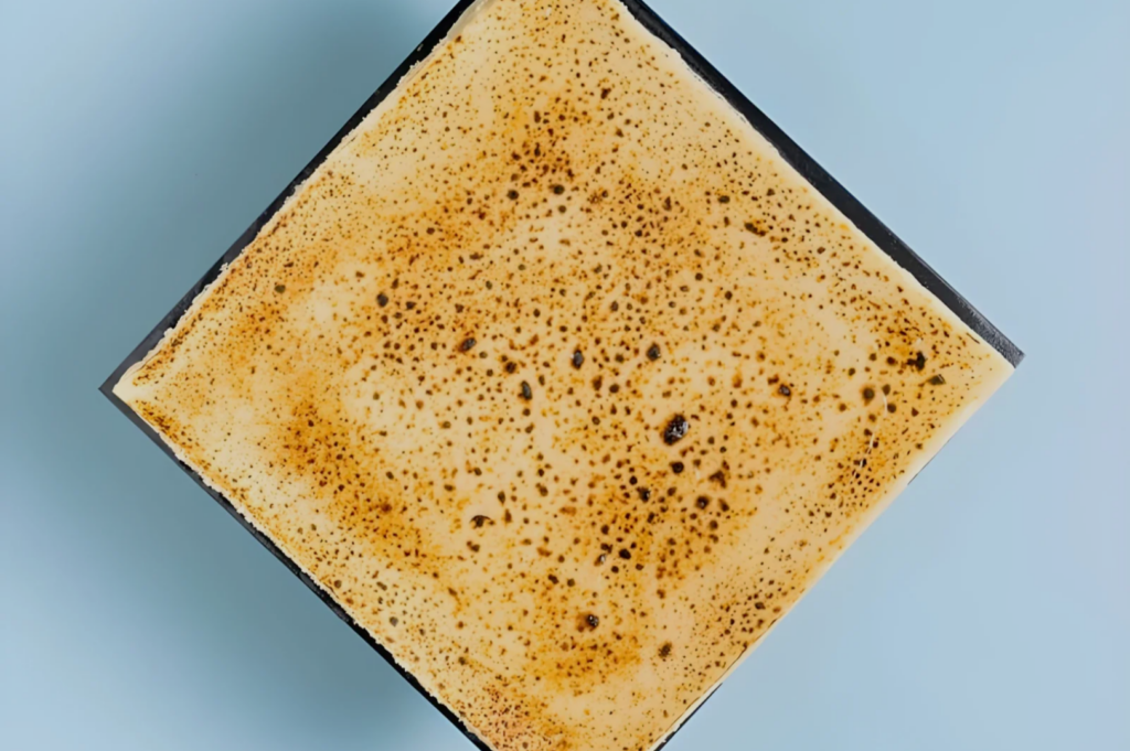 A square dessert with a golden, caramelized top, resembling crème brûlée, sits on a light blue background, conveying a sense of elegance and sweetness.