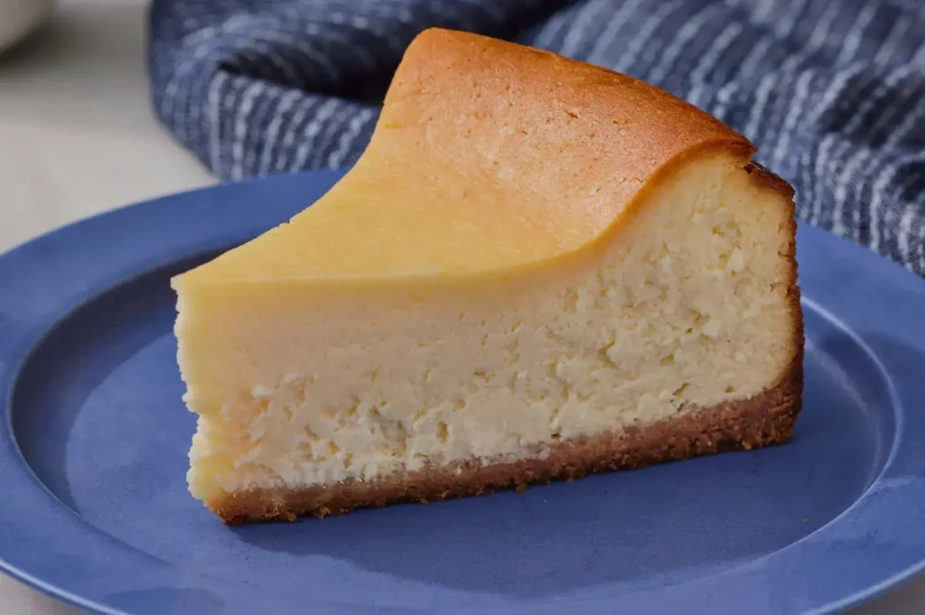 A thick slice of creamy cheesecake with a golden-brown top and crumb crust rests on a blue plate, inviting and freshly served.