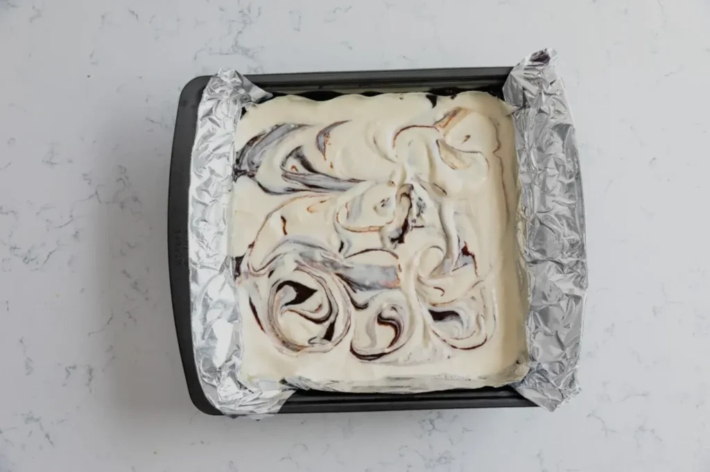 Overhead view of a foil-lined baking pan filled with creamy dessert batter swirled with chocolate, ready to bake; the homemade treat looks rich and inviting.