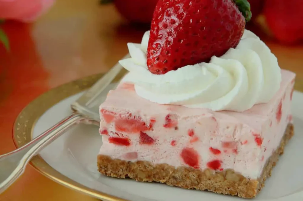A creamy strawberry cheesecake bar topped with whipped cream and a fresh berry sits on a plate with a fork, inviting and indulgent.