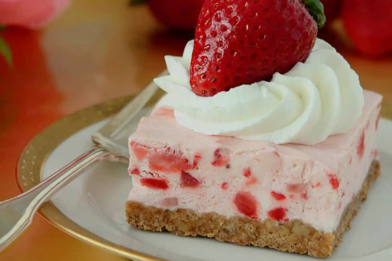 A creamy strawberry cheesecake bar topped with whipped cream and a fresh berry sits on a plate with a fork, inviting and indulgent.