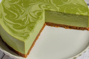 A slice of pale green, marbled cheesecake with a golden crumb crust is cut from a round cake, creating a fresh, elegant dessert presentation.