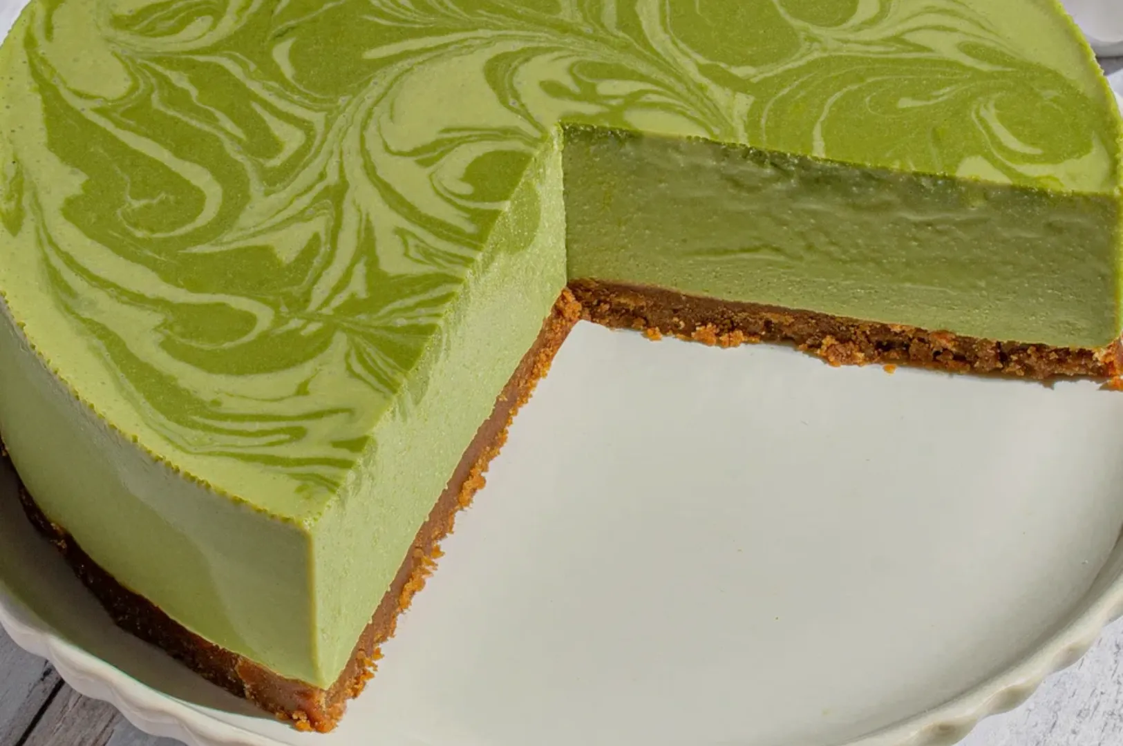 A slice of pale green, marbled cheesecake with a golden crumb crust is cut from a round cake, creating a fresh, elegant dessert presentation.