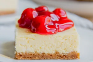 A creamy cheesecake bar with a golden crumb crust is topped with glossy red cherry filling, photographed close-up for an inviting, indulgent dessert look.