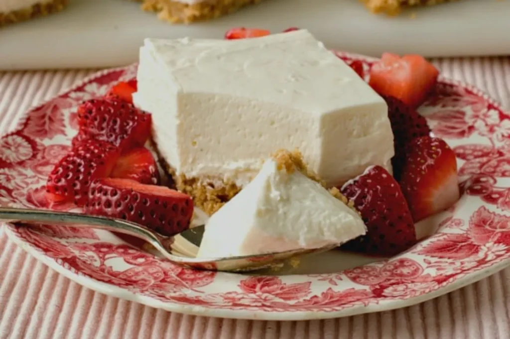 A creamy cheesecake slice surrounded by fresh strawberries on a red floral plate, with a fork ready to serve; the dessert looks rich and inviting.