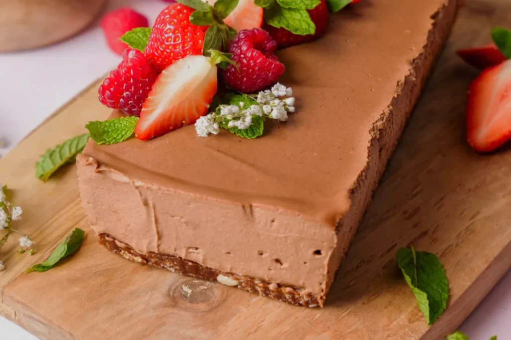 A smooth rectangular chocolate mousse cake on a wooden board, topped with strawberries, raspberries, mint, and tiny white flowers; rich, inviting dessert.