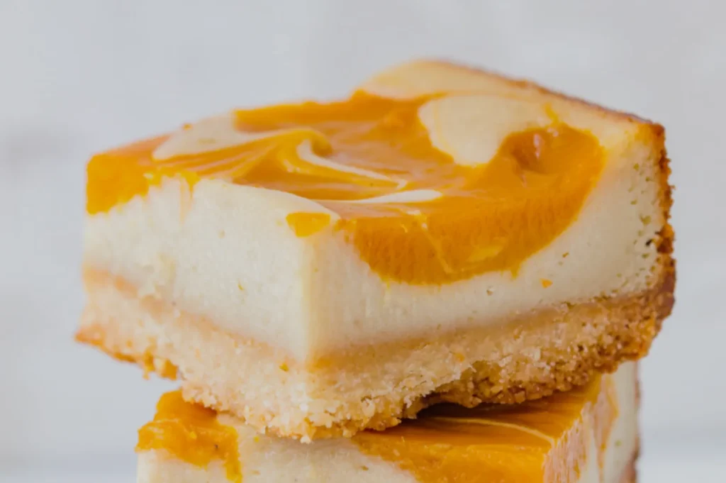 A rich slice of creamy orange-swirl cheesecake with a golden crumb crust rests atop another slice, photographed close-up against a pale background.