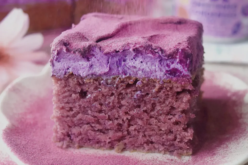 A thick slice of purple berry cake with a creamy lavender filling and soft crumb sits on a plate dusted with pink powder, creating a cozy, sweet mood.