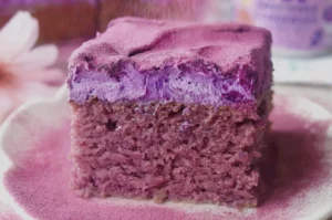 A thick slice of purple berry cake with a creamy lavender filling and soft crumb sits on a plate dusted with pink powder, creating a cozy, sweet mood.