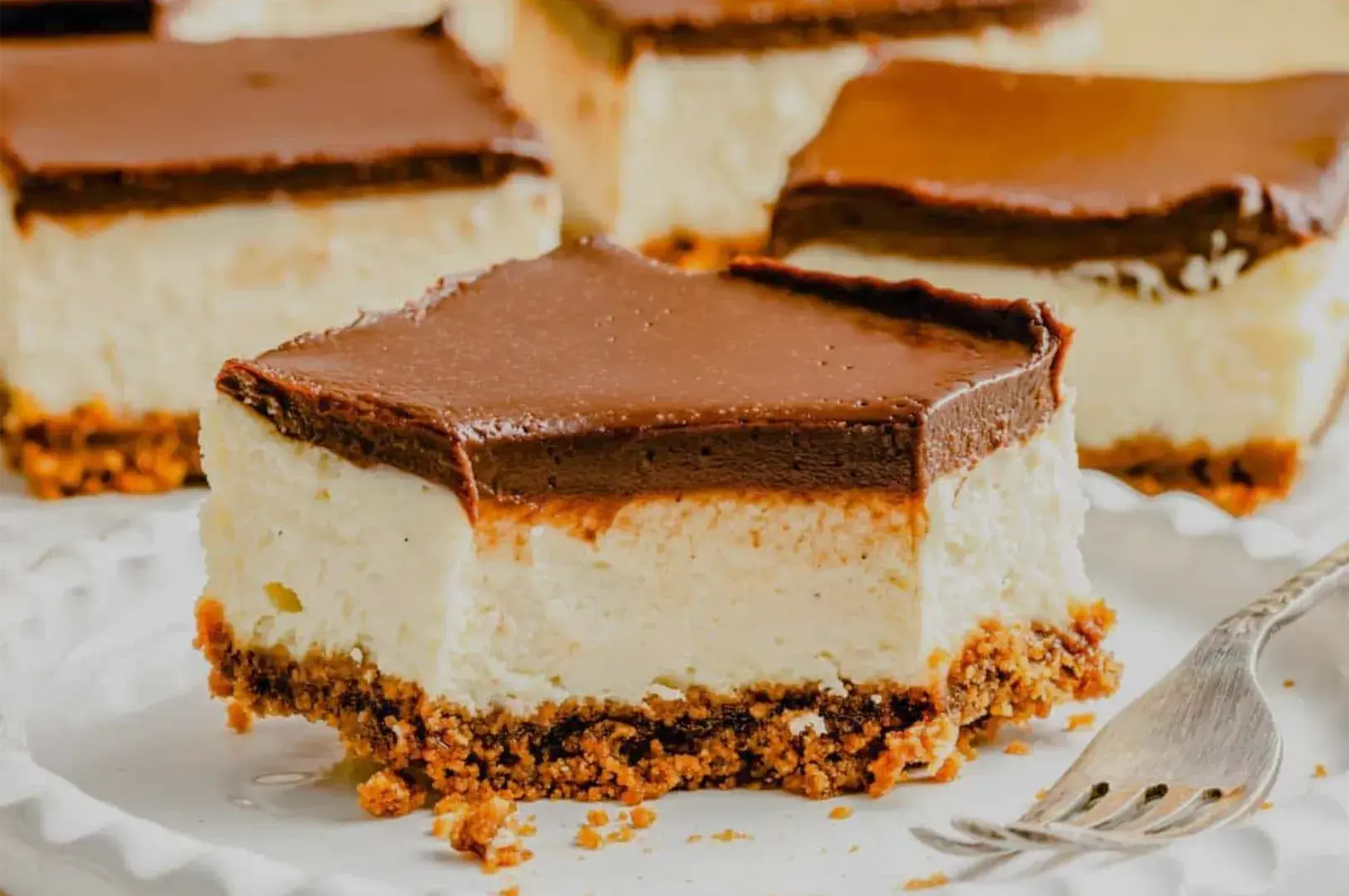 A creamy layered cheesecake square with chocolate topping and crumb crust sits on a plate, with more slices behind it and a fork nearby; the scene feels warm and inviting.